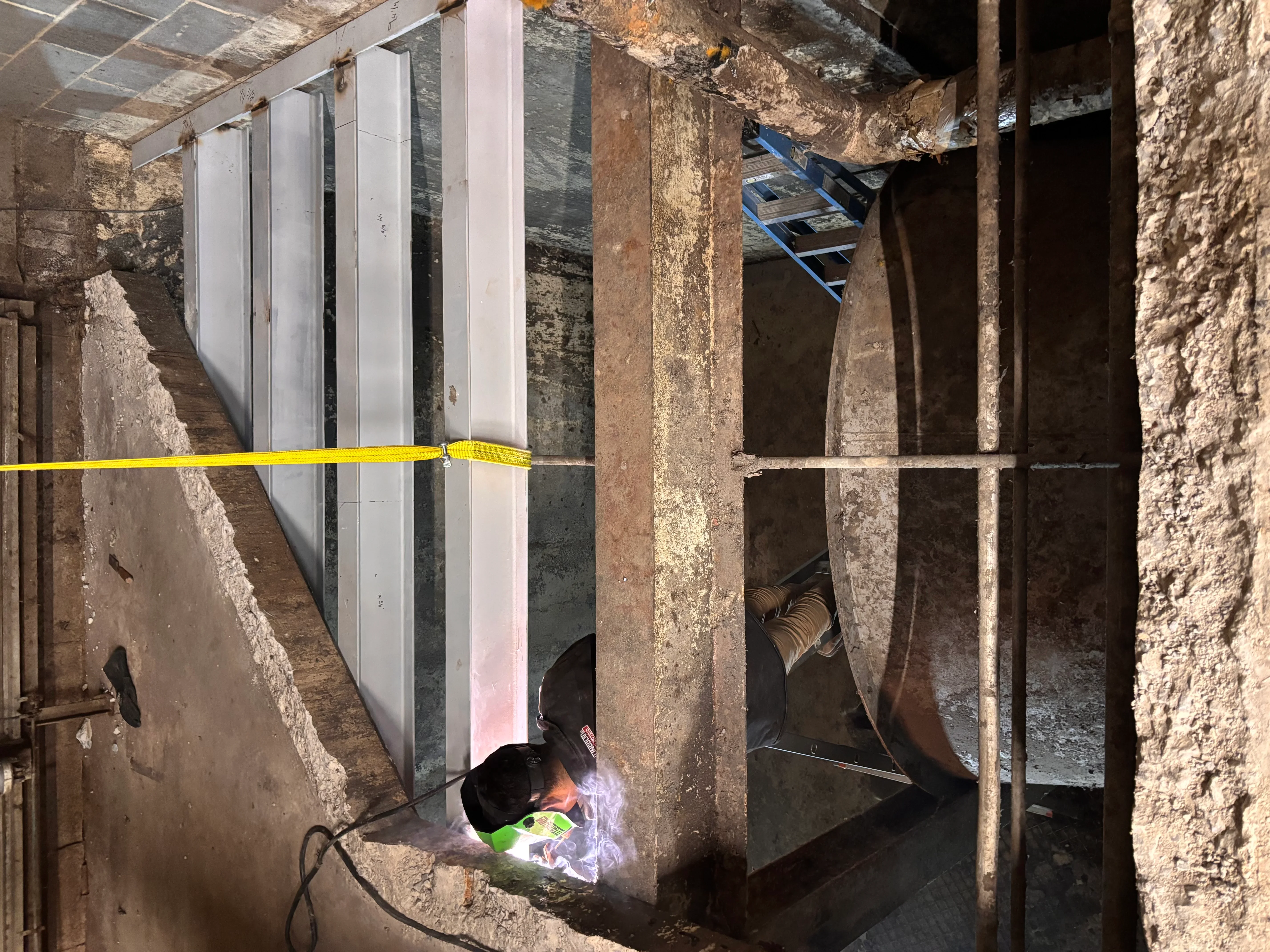 Welder joining structural steel beams in basement with safety tape and equipment visible